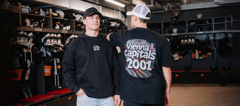Zwei Männer stehen in einer Eishockey-Umkleidekabine; der eine schaut nach vorne und trägt einen schwarzen Kapuzenpulli und eine Mütze, der andere schaut nach hinten und trägt ein schwarzes Vienna Capitals 2001 T-Shirt und eine weiße Trucker-Mütze.