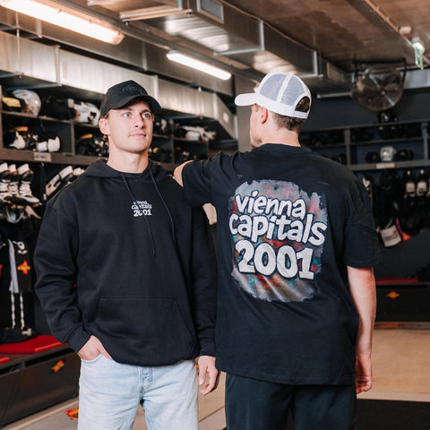 Zwei Männer stehen in einer Eishockey-Umkleidekabine; der eine schaut nach vorne und trägt einen schwarzen Kapuzenpulli und eine Mütze, der andere schaut nach hinten und trägt ein schwarzes Vienna Capitals 2001 T-Shirt und eine weiße Trucker-Mütze.