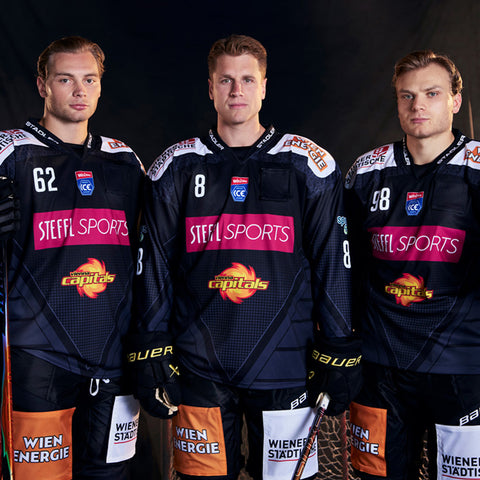 Drei männliche Eishockeyspieler in schwarzen Uniformen der Vienna Capitals posieren gemeinsam mit ihren Schlägern vor einem dunklen Hintergrund.