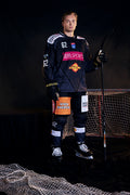 Eishockeyspieler im dunkelblauen Vienna-Capitals-Trikot mit Nummer 62 steht mit Schläger vor einem Eishockeytor.